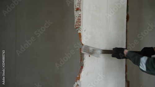 Close-up view of construction worker wearing protective mask and gloves using heavy sledgehammer to break through brick wall plaster during home renovation project