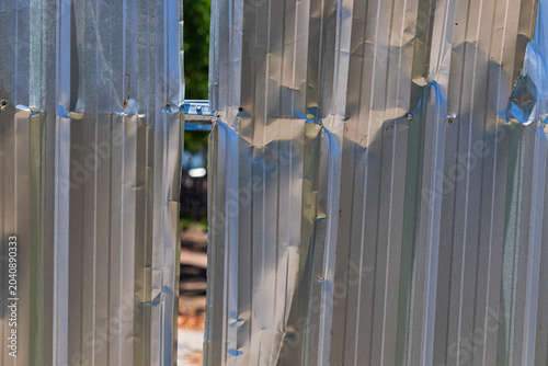 Fotografija Close-up of a damaged and dented corrugated metal sheet fence with a vertical gap revealing a blurred outdoor background