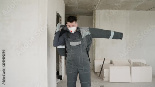 A male construction worker in a grey uniform puts on a jacket and removes a dust mask in an unfinished building interior