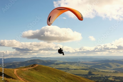 Solo paraglider flies high above rolling green hills and distant mountains. Person enjoys freedom in the sky with clouds and clear blue sky. Active leisure.