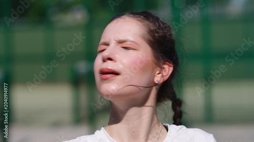 Tired teenage athlete resting and drinking water from a bottle after an intense match on a sunny day. She is sweating and exhausted
