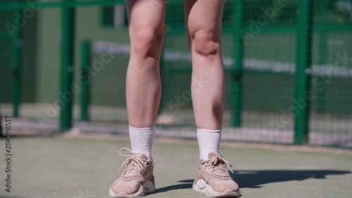 Low section shot of a woman holding a racket and bouncing a padel ball with her feet on a green court. Warm-up routine before the match