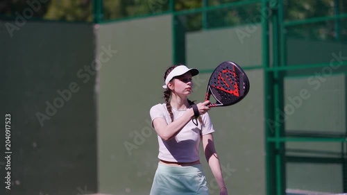 Focused young woman in sportswear serving the ball with a padel racket. Athletic girl wearing a visor, training on a green court