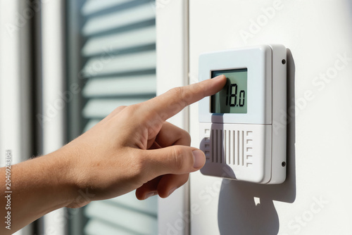 Hand adjusts digital thermostat setting lower for energy saving. Person controls indoor temperature reducing home heating costs for better economy. Adjusting warmth efficiency for winter.