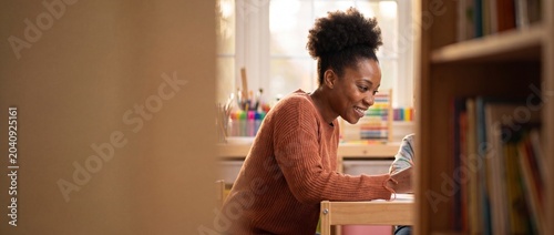 Black teacher helping child drawing in classroom, African American mother homeschooling kid at desk, warm educational classroom background, happy preschool tutor supporting early education.