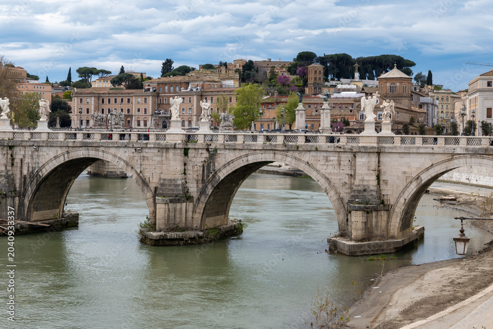 Fototapeta premium View on stone-arched bridge on Tiber river in Rome, balustrade, marble statues, cloudy sky, ochre and terracotta buildings, domes and umbrella pines rise on gently sloping Vatican hill