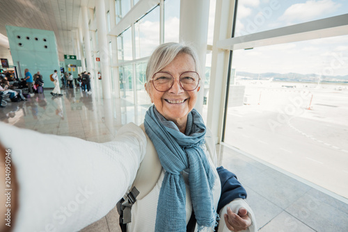 Schilderij op canvas Senior woman is happily taking a selfie while waiting in a bright, modern airpor