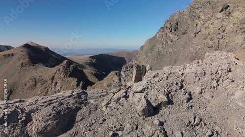 Drone footage captures mountain goats bounding along a barren ridge through harsh alpine terrain, with dramatic rocky cliffs, steep slopes and clear blue sky above distant mountain peaks