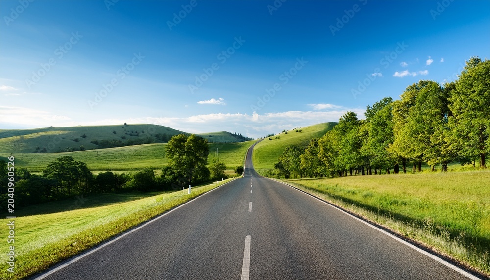 Fototapeta premium long empty road stretches into distance surrounded by lush green trees and rolling hills under clear blue sky serene landscape evokes sense of calm and adventure