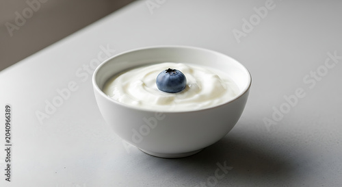 A single blueberry floating in a white bowl of yogurt on a gray surface