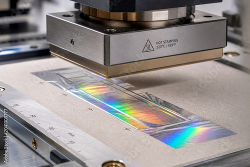 A hot stamping machine works on a piece of paper using a rainbow foil. The setup shows the machine applying heat to transfer the foil onto the surface. This takes place in a factory during the day.