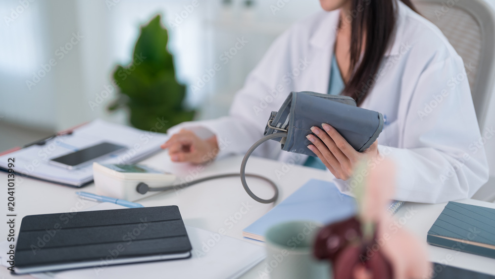 Fototapeta premium Asian adult female doctor measuring blood pressure with device at medical clinic office desk for health checkup examination cardiovascular hypertension