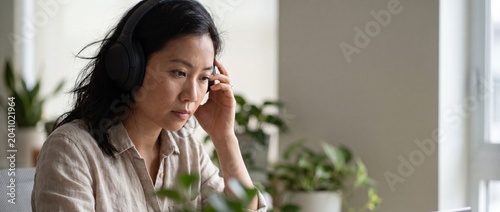 Asian woman wearing headphones working online in home office, looking focused and serious while concentrating on computer screen laptop, remote business employee studying or listening to podcast