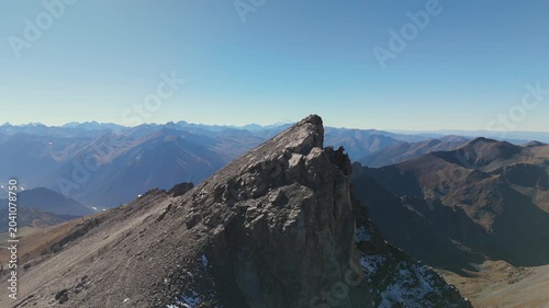 Aerial drone video flying above a rocky mountain summit in high alpine terrain, revealing sharp ridgelines, distant peaks and vast wilderness beneath clear blue sky in bright autumn daylight