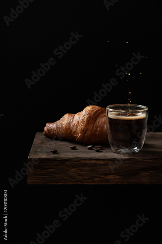 Espresso coffee splash with fresh croissant on rustic wooden table, dark background and copy space