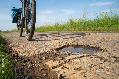 Cyclist rides past a pothole on a road with grass on the side during a sunny day in an open field