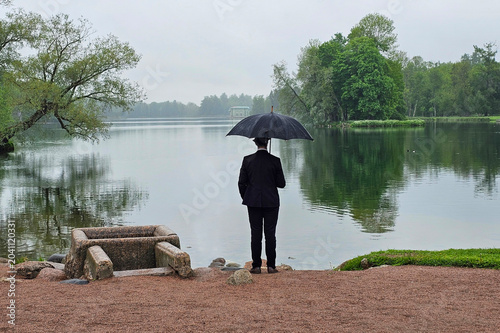 Rainy day, lone figure stands by calm lake, view from the back.