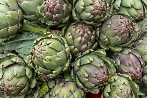A close-up of a pile of fresh whole globe artichokes with green and purple-tinged leaves on display at a market stall