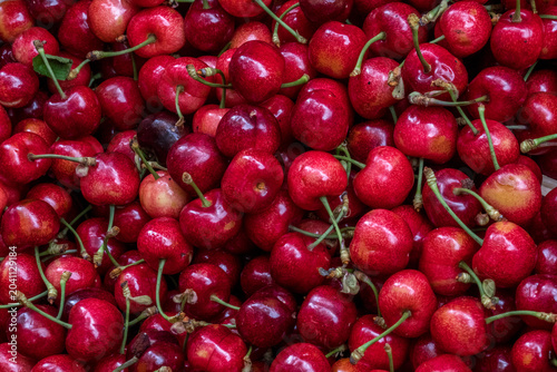 A vibrant close-up of a large pile of fresh ripe red cherries with green stems, glistening with moisture at a market stall