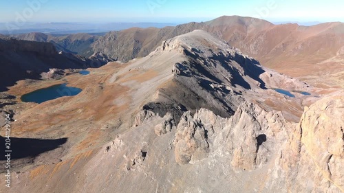 Cinematic drone footage gliding above high alpine mountains, revealing blue lakes, sharp rocky ridges, barren slopes and vast remote wilderness under clear blue sky in bright autumn daylight