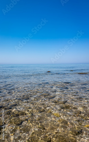 Crystal clear shallow sea water with pebbles and blue sky horizon. A vertical, serene view of shallow, transparent water with submerged pebbles on the seabed. Perfect for themes of summer, vacation.