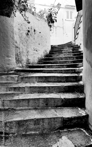A moody, high-contrast black and white photograph of an ancient stone stairway winding up through a narrow alleyway between old buildings. The scene evokes a sense of history, mystery, and exploration