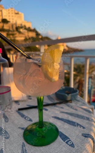 Refreshing cocktail with lemon slice at sunset. A vertical close-up shot of a refreshing cocktail garnished with a lemon slice, served in a glass with a green stem.