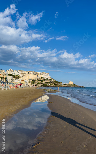 Historic coastal town overlooking a sandy beach. A vertical view showing a historic Mediterranean town perched on a cliff above a sandy beach. A tide pool in the foreground mirrors the sky and town