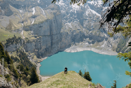 Couple Overlooking Alpine Lake. A serene moment captured in the Swiss Alps: a bald man and his companion stand on a grassy cliff edge. Beautiful landscapes in the Swiss Alps, Oeschinensee