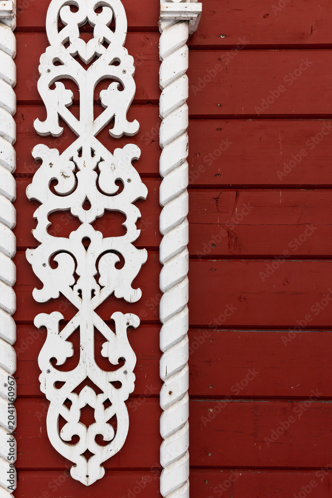 Fototapeta premium Carved white wooden trim with ornate scrollwork flanked by twisted columns on a red painted wooden wall, offering rustic, traditional architectural detail and vintage charm for Russian exterior design