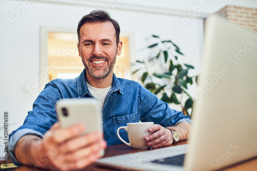 Man smiling with smartphone and coffee at home desk