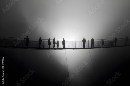 Silhouetted figures stand on a suspension bridge shrouded in dense fog, creating a dramatic, high-contrast black and white scene.