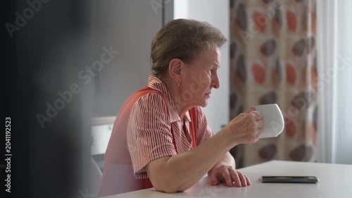 An elderly sad pensive woman in the kitchen drinking tea