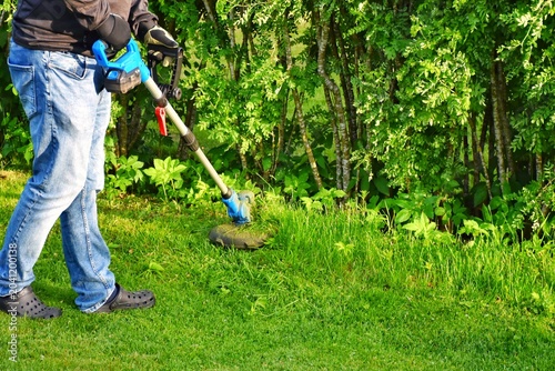 Fotografie A man trims the grass near a bush with a cordless trimmer.