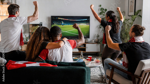 Enthusiastic multi-ethnic friends cheering and celebrating a goal while watching a professional soccer match on a large TV at home in a cozy living room.