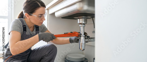 A woman is fixing the plumbing under a kitchen sink. She uses a wrench to secure pipes and wears gloves for protection. This happens during daylight hours in a home kitchen
