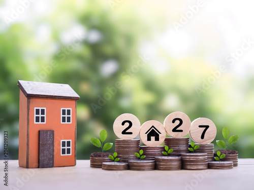 Miniature house, Wooden blocks with the number 2027 and plants growing up on stack of coins. The concept of saving money for house, Property investment, House mortgage, Real estate in year 2027