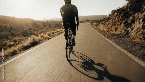 Tracking shot of cyclist riding along winding desert road with long shadow at sunset