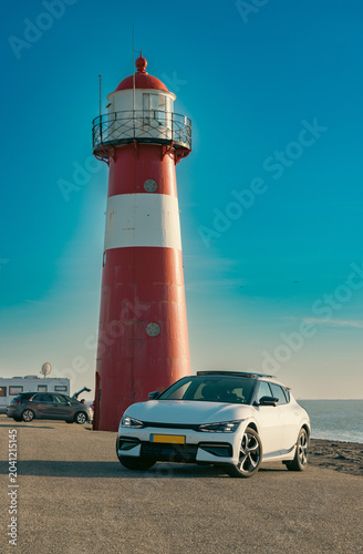 Modern Electric Car Parked Near Red and White Lighthouse on Dutch Coast