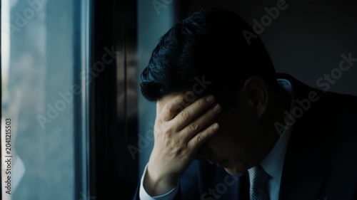 Slow dolly in of businessman looking out window then closing eyes and covering face in office interior during daytime