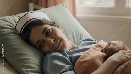 Slow tracking shot of nurse resting and smiling while lying in hospital bed by window in quiet ward during morning light