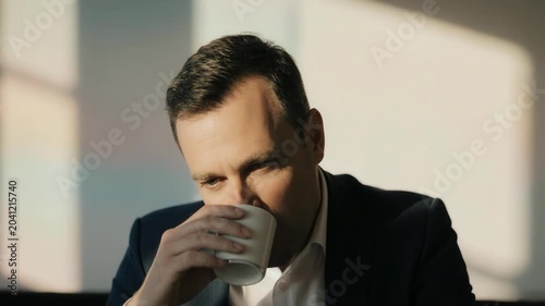 Static shot of businessman smiling, blinking and sipping coffee in sunlit office interior near window