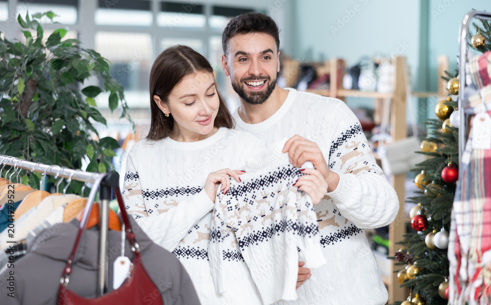 Obraz premium Married couple choose sweaters against background of Christmas tree in a clothing store