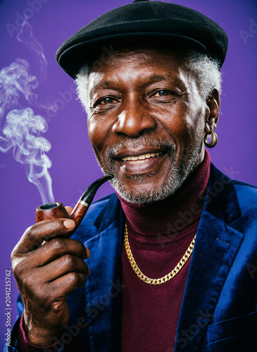 An elderly African American man shining a brass lantern and holding a smoking pipe while posing against a purple background
