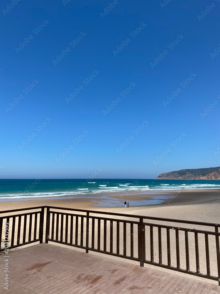 Fototapeta premium Wooden terrace overlooks wide sandy beach with people walking near Atlantic Ocean waves on sunny day in Portugal