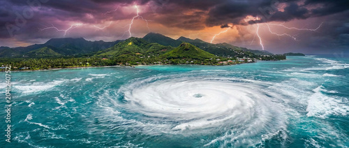 Dramatic aerial view of a giant ocean whirlpool forming near a tropical island under lightning filled storm clouds. Extreme weather, climate change and natural disaster concept.