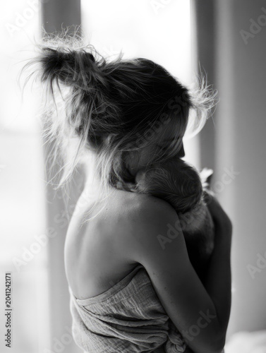 Postpartum mother hugging newborn back view messy hair bun tired posture natural light close up black and white breastfeeding top soft fabric intimate family moment