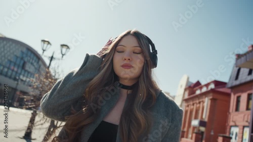 Sunlit city street portrait of woman in coat and beanie, headphones on, looking up at glass market and redbrick buildings peaceful expression, cinematic wideangle, soft cool tones, gentle breeze