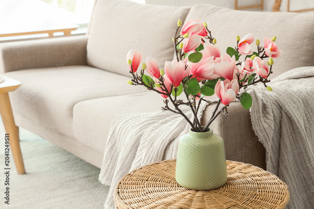 Obraz premium Vase with beautiful magnolia flowers on wicker table near sofa in living room, closeup