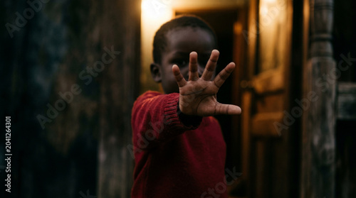 African child holding hand out in stop gesture, young boy in red sweater, cinematic portrait of social issues